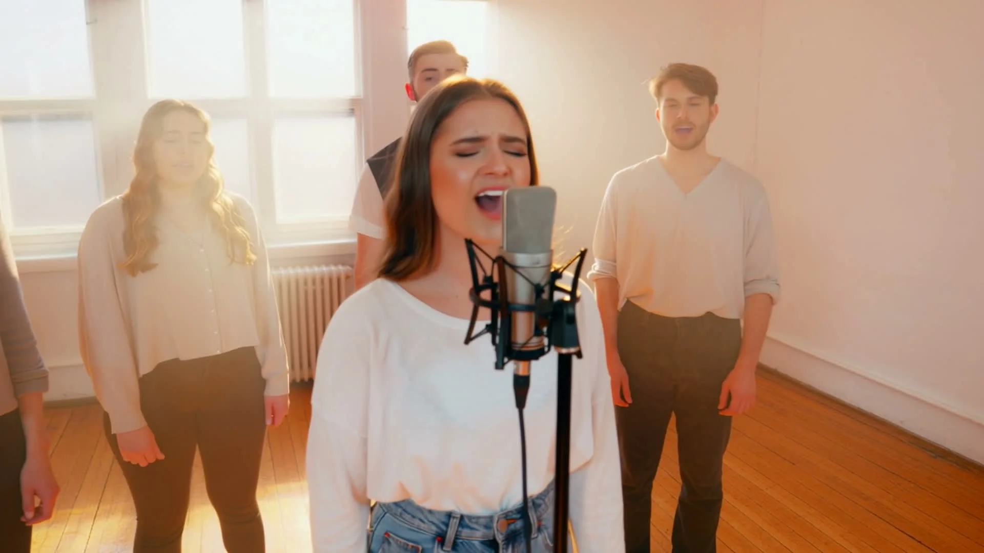 A young woman sings passionately into a professional studio microphone with three backup singers behind her, in a bright sunlit room with large windows, wooden floors and warm golden-hour glow
