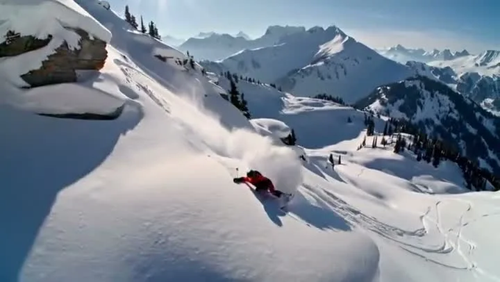 A skier in a red jacket carves through deep powder snow on a steep slope, creating a dramatic white spray against majestic snow-covered peaks under a clear blue sky